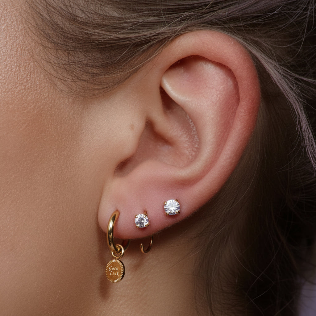 Close-up of an ear wearing Gold-plated stainless steel huggie hoop earrings with an engraved Good Luck disc charm – naušnice