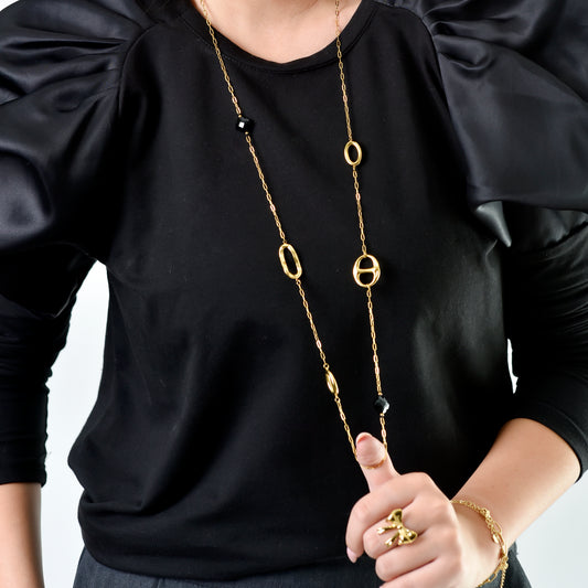 Person wearing a black shirt with a long gold necklace and accessories on a plain background